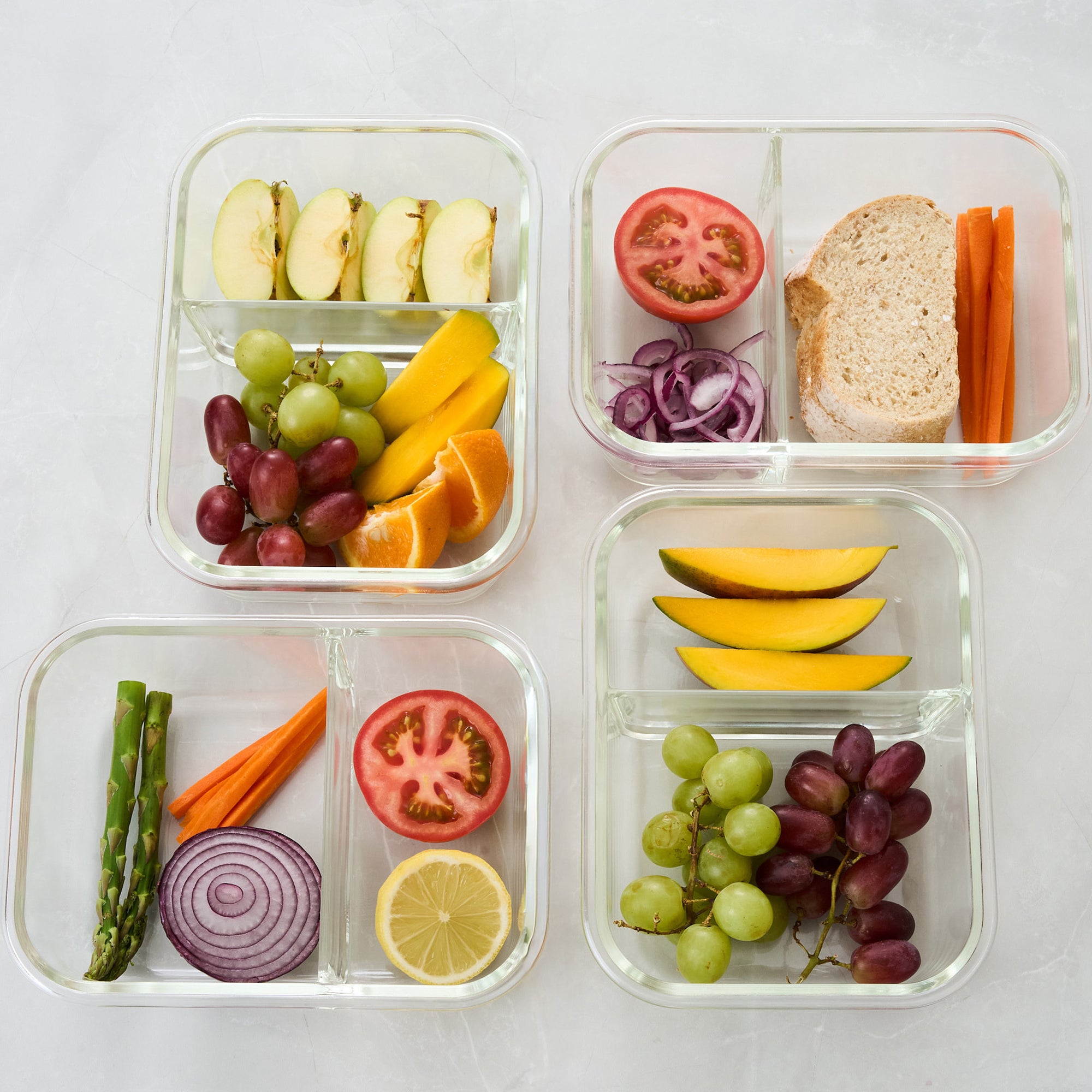 Overhead view of four open Storinity divided glass containers filled with assorted fruit, vegetables and a sandwich