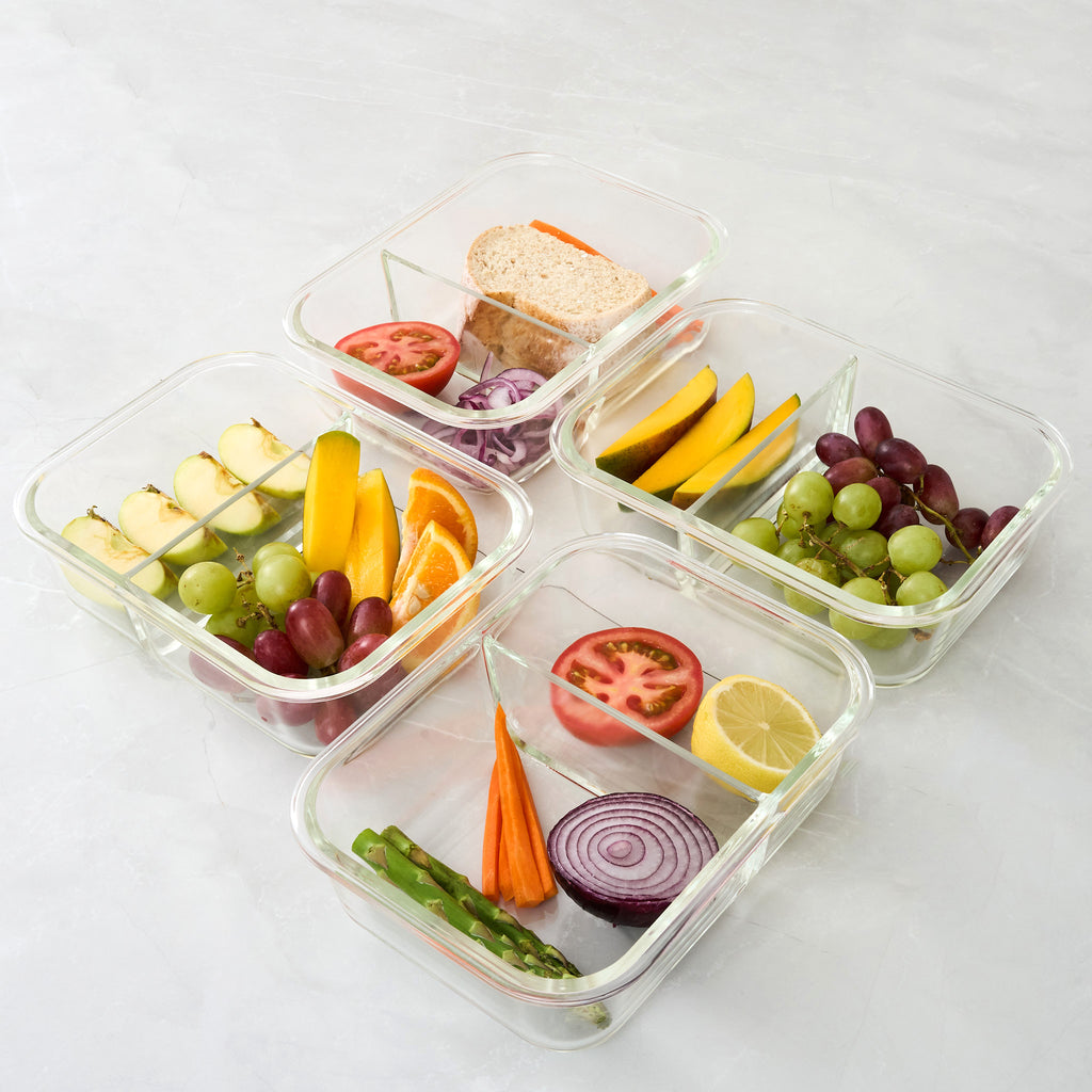 Five clear glass compartment lunch containers arranged on a light marble-like surface, filled with sliced vegetables, grapes, orange, apple, and a sandwich.