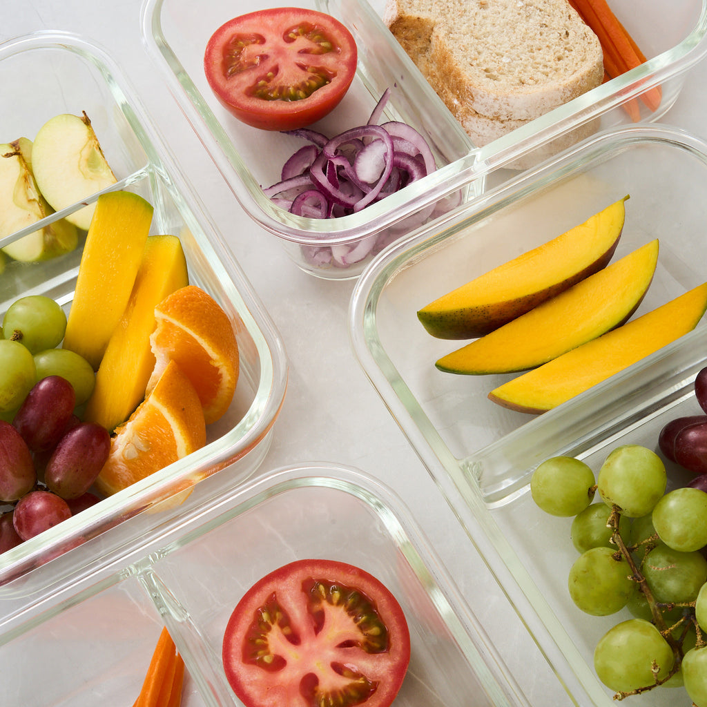 Clear glass compartment food storage containers filled with sliced fruit, grapes, orange wedges, red onion, tomato, and a sandwich slice on a white surface.