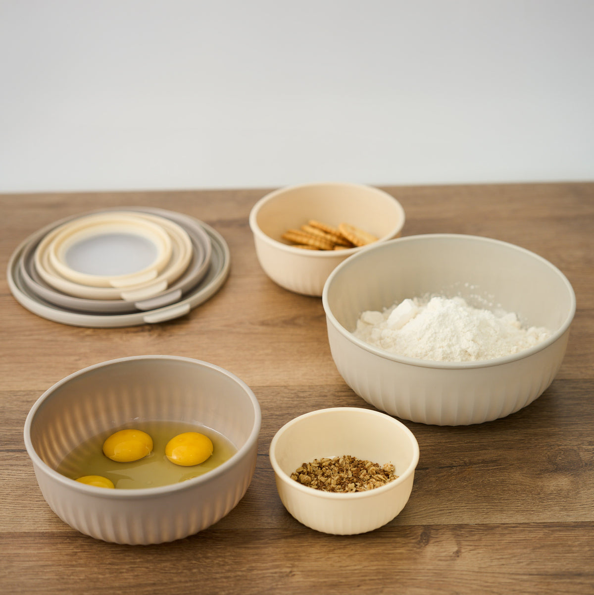 Storinity nesting bowls filled with crackers, flour, eggs and oats on a wooden countertop, with the lids stacked beside them