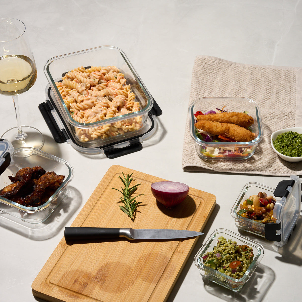 Storinity glass containers filled with pasta salad, chicken wings, crumbed fish and bean salad beside a glass of wine and cutting board