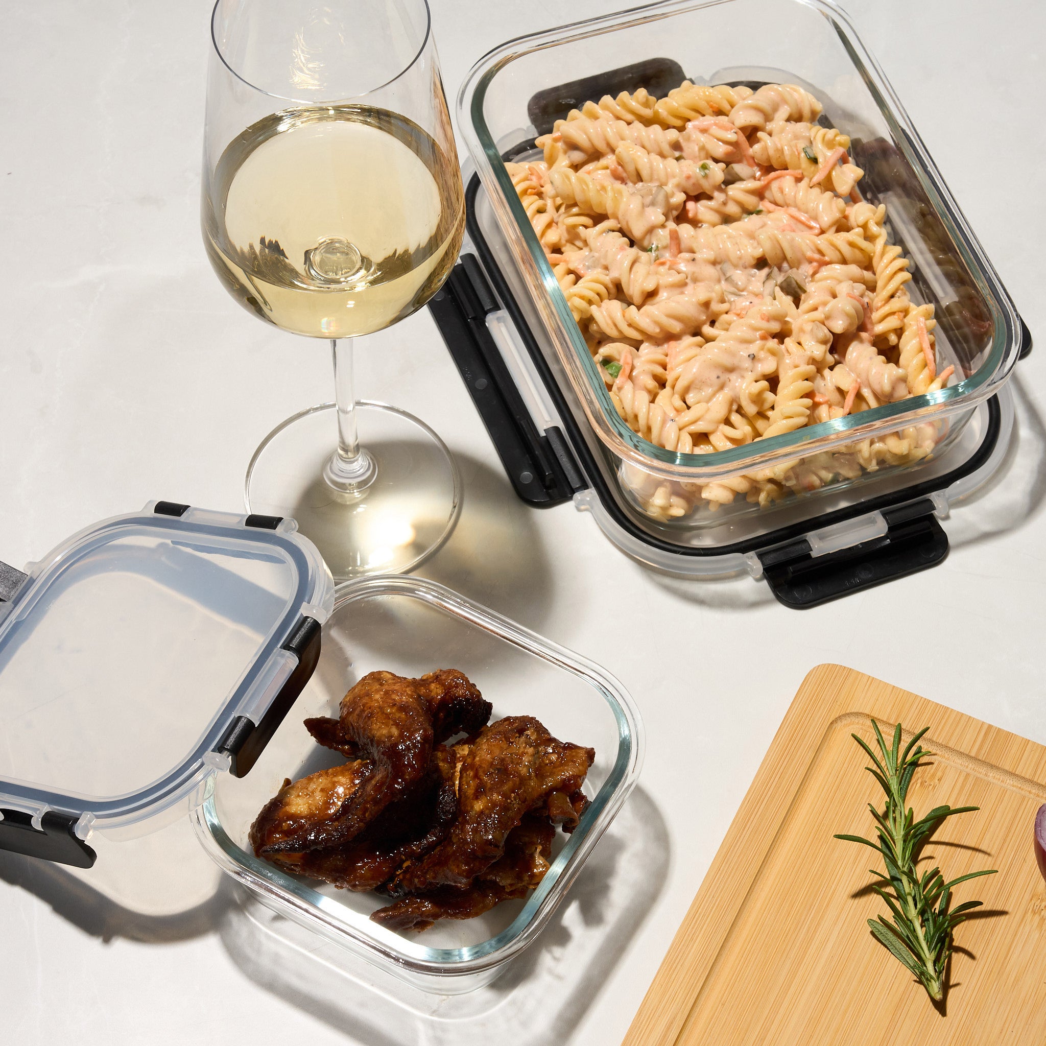 Storinity glass containers filled with pasta salad and glazed chicken wings beside a glass of white wine and rosemary