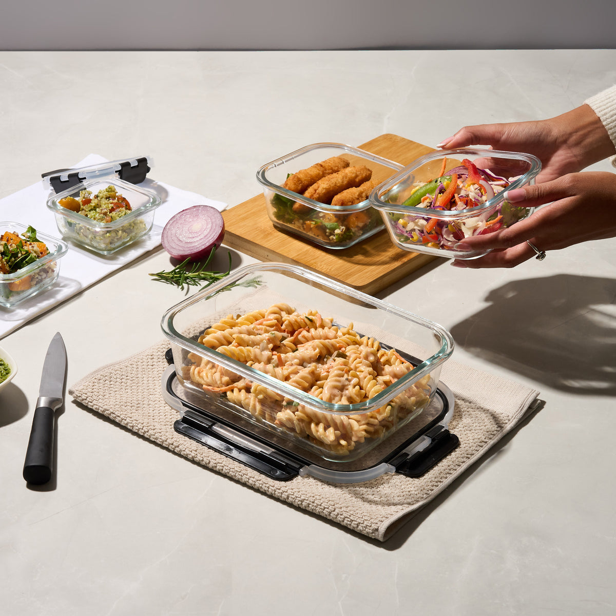 Hands reaching for a Storinity glass container of coleslaw beside containers of pasta salad and crumbed fish on a kitchen counter