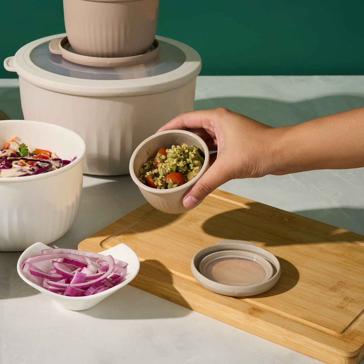 Hand holding a small Storinity nesting bowl of rice and pesto salad beside larger bowls of coleslaw and sliced red onion