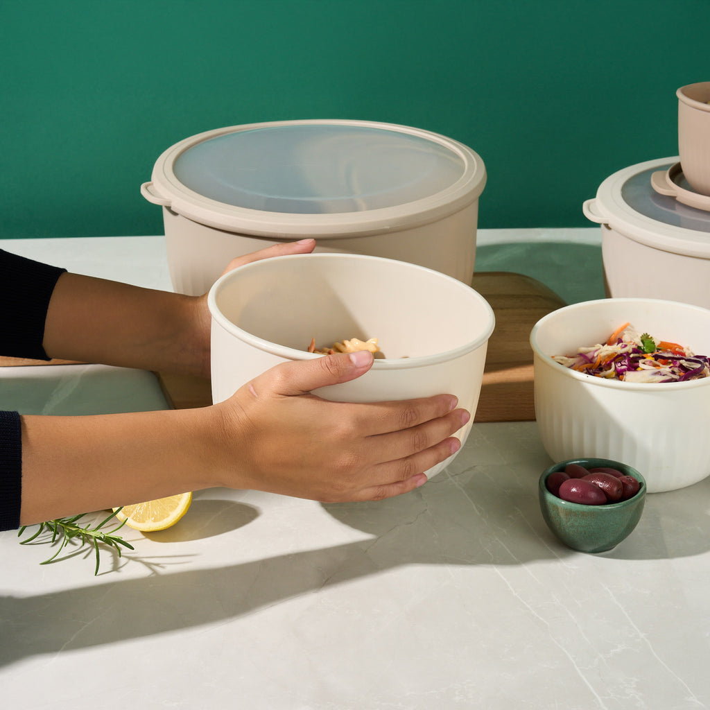 Hands holding a Storinity nesting bowl of pasta salad beside lidded bowls, coleslaw, olives and lemon on a green backdrop