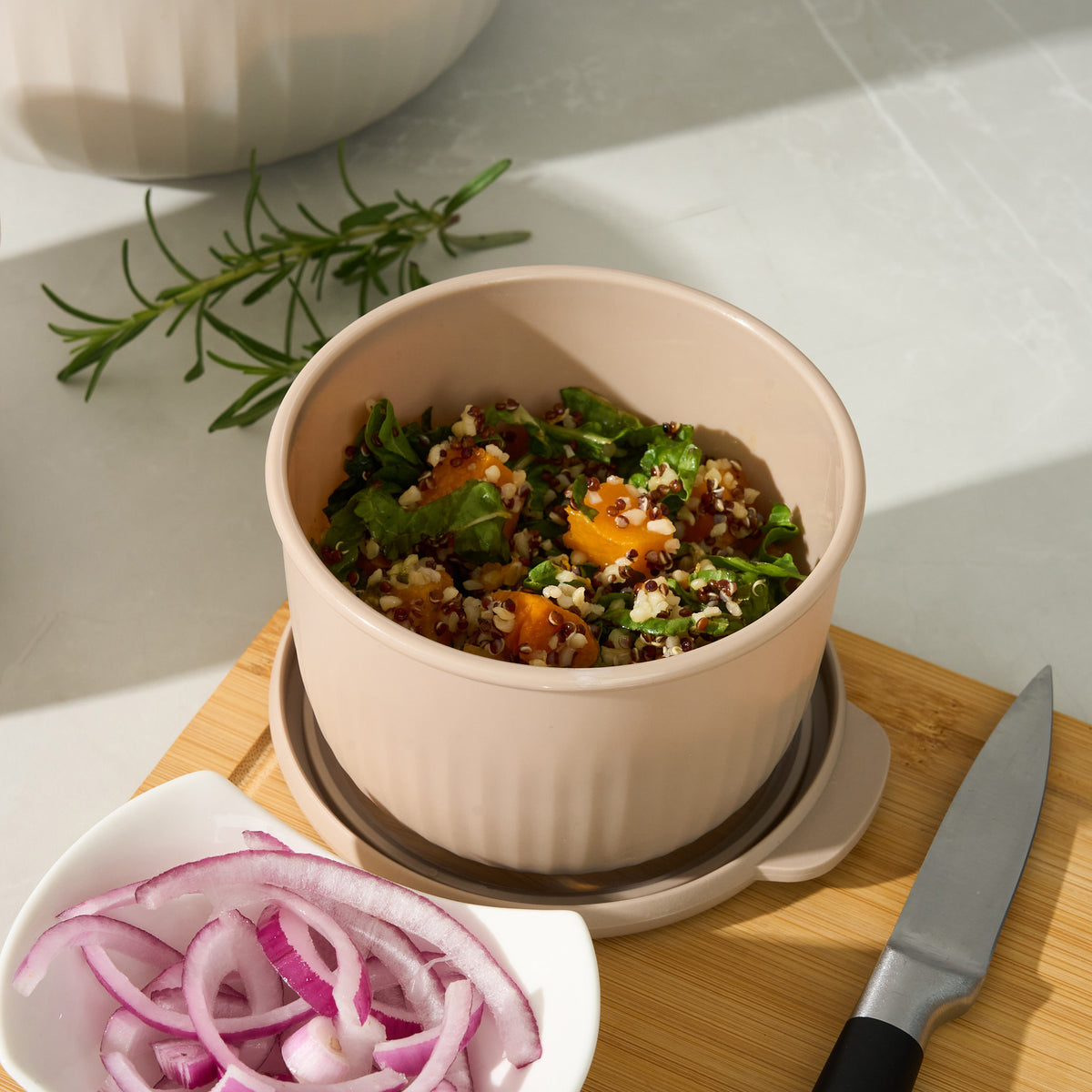 Storinity nesting bowl filled with quinoa, pumpkin and greens on a cutting board with rosemary, red onion and a knife