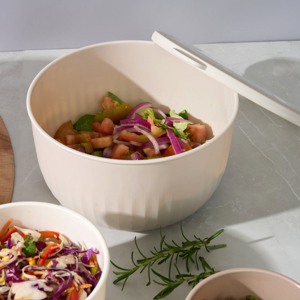 Open Storinity nesting bowl of tomato and red onion salad with the lid propped open, beside a bowl of coleslaw and rosemary