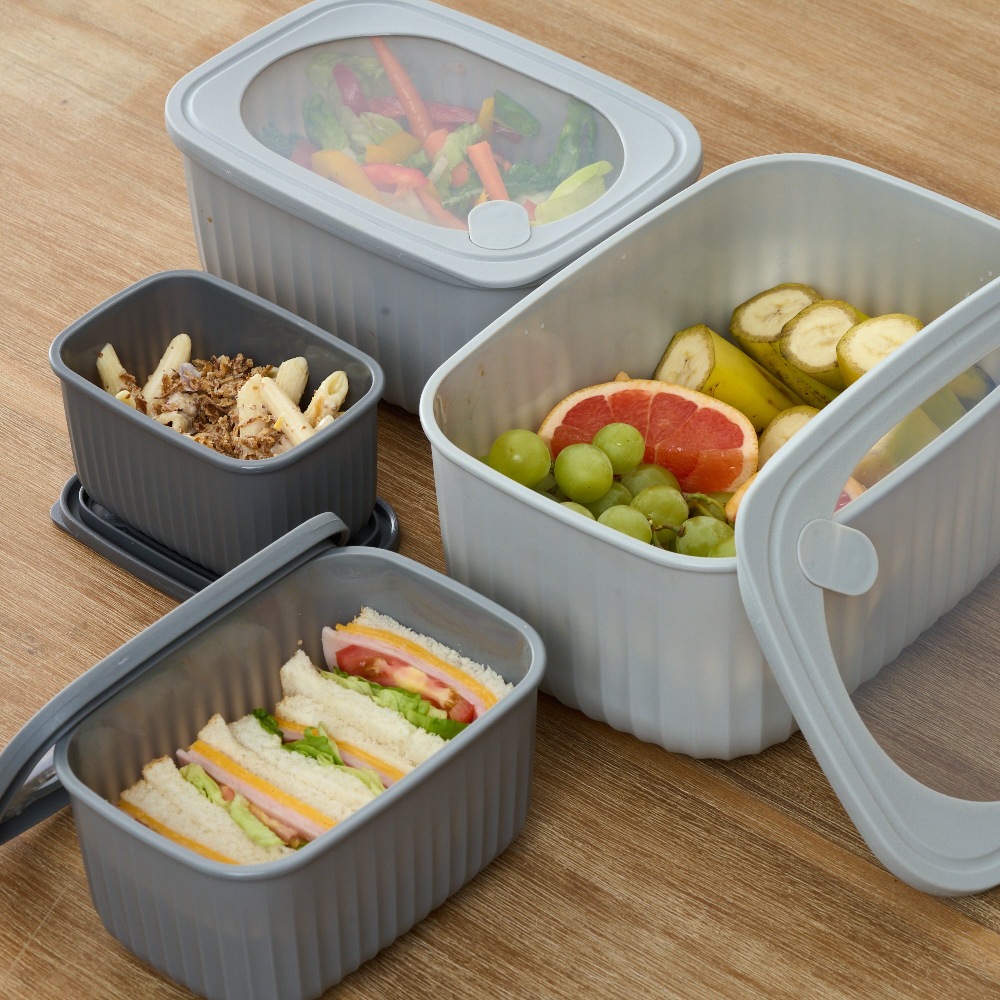 Grey Storinity rectangular nesting bowls filled with salad, fruit, pasta and sandwiches on a wooden table